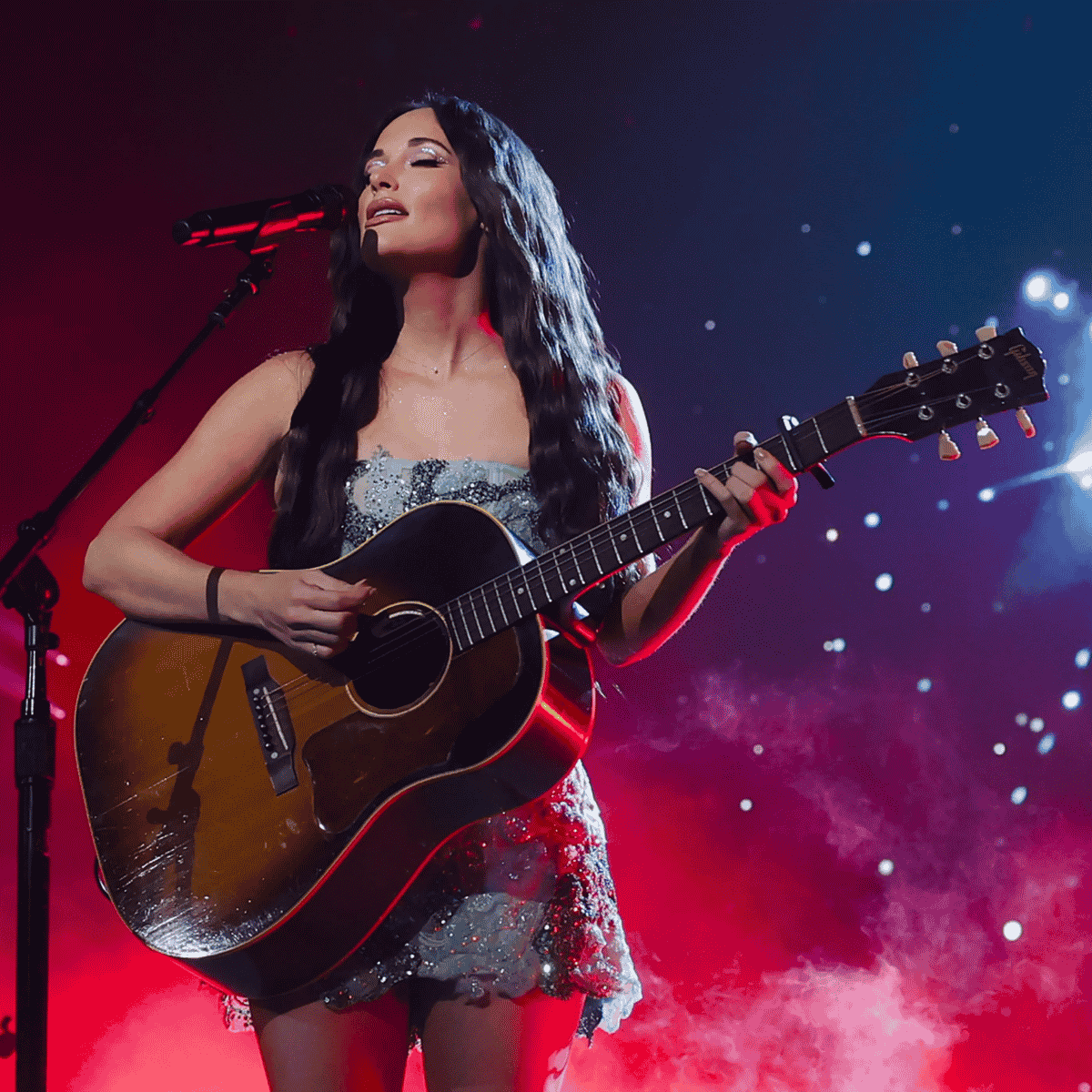Kacey Musgraves akusztikus gitárral koncert közben, vörös és kék fényekben