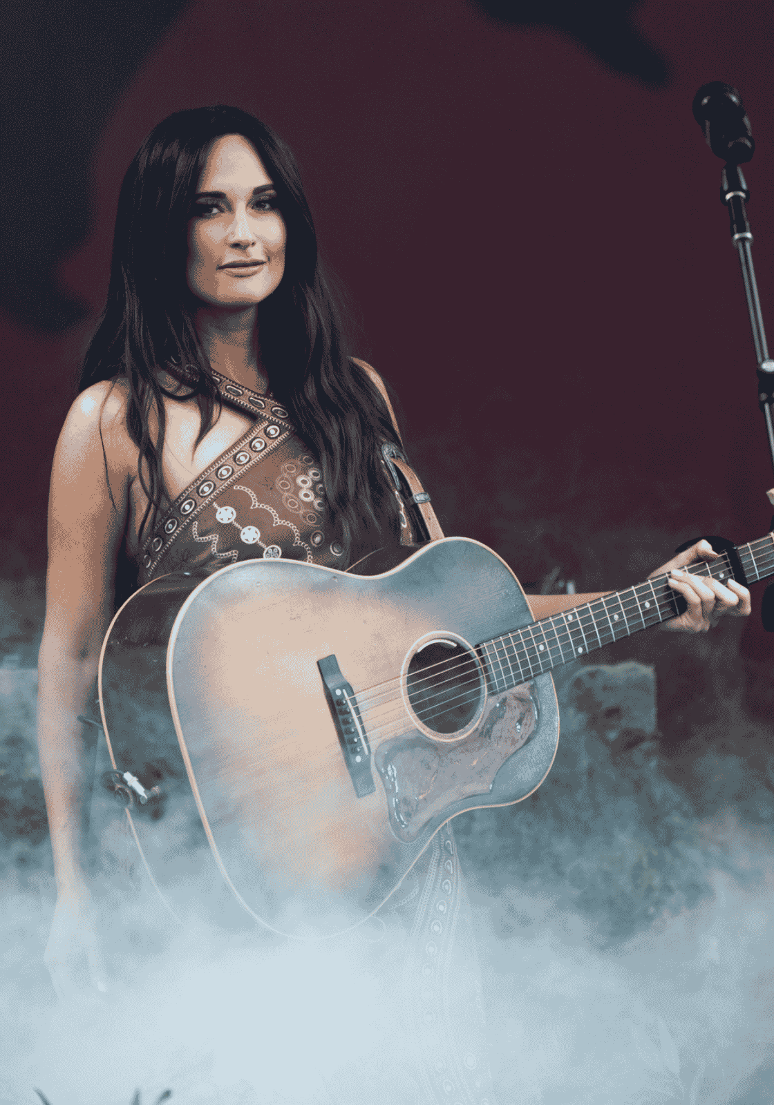 Kacey Musgraves akusztikus gitárral, bordó színpadi háttér és köd előtt